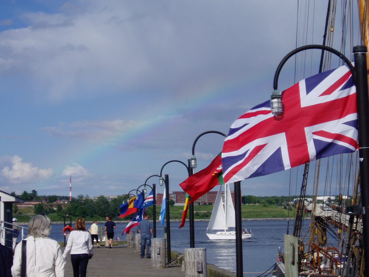 Visitors strolling the Halifax waterfront boardwalk past tall ships and flags on a sunny day