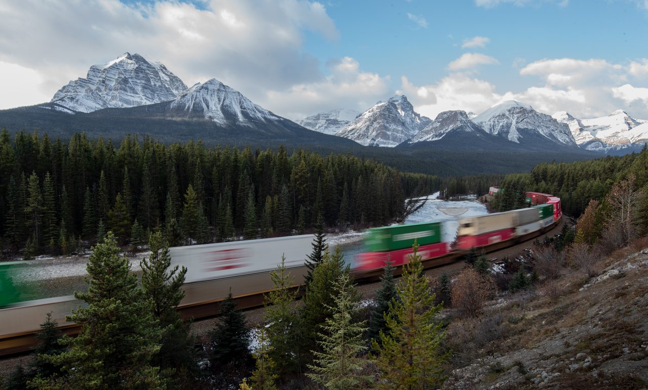 VIA Rail's The Canadian train curving along the river at Morant's Curve with Rocky Mountain peaks behind