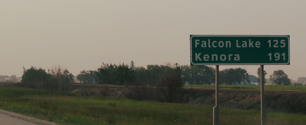 Long straight stretch of the Trans-Canada Highway through flat prairie with a green sign showing 191 km to Kenora, Ontario