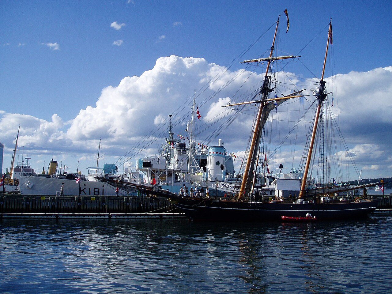 Tall ship and naval vessels docked along the Halifax waterfront boardwalk under a blue sky